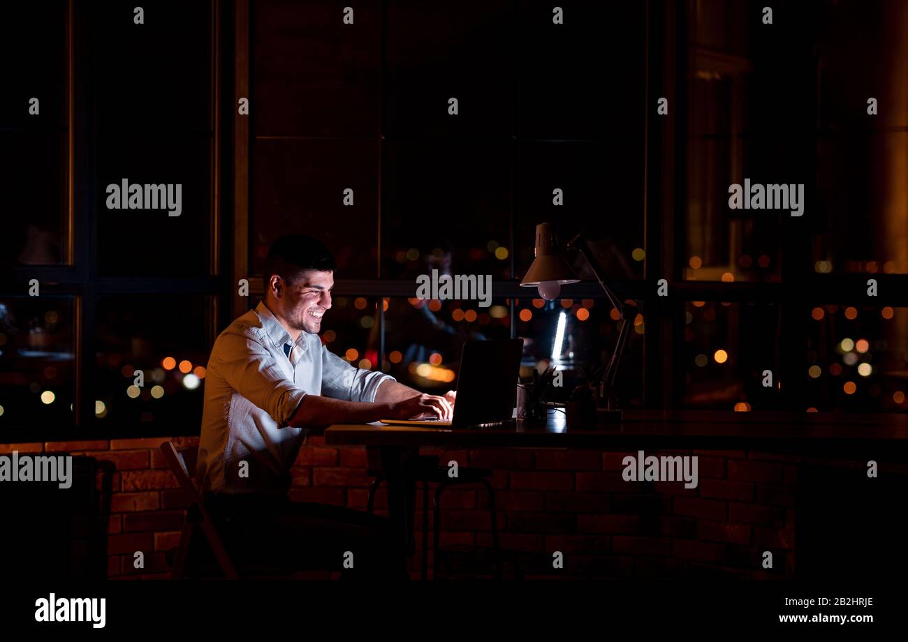 Gioioso Uomo Che Lavora Su Laptop Online Seduto Di Notte In Interni Foto Stock