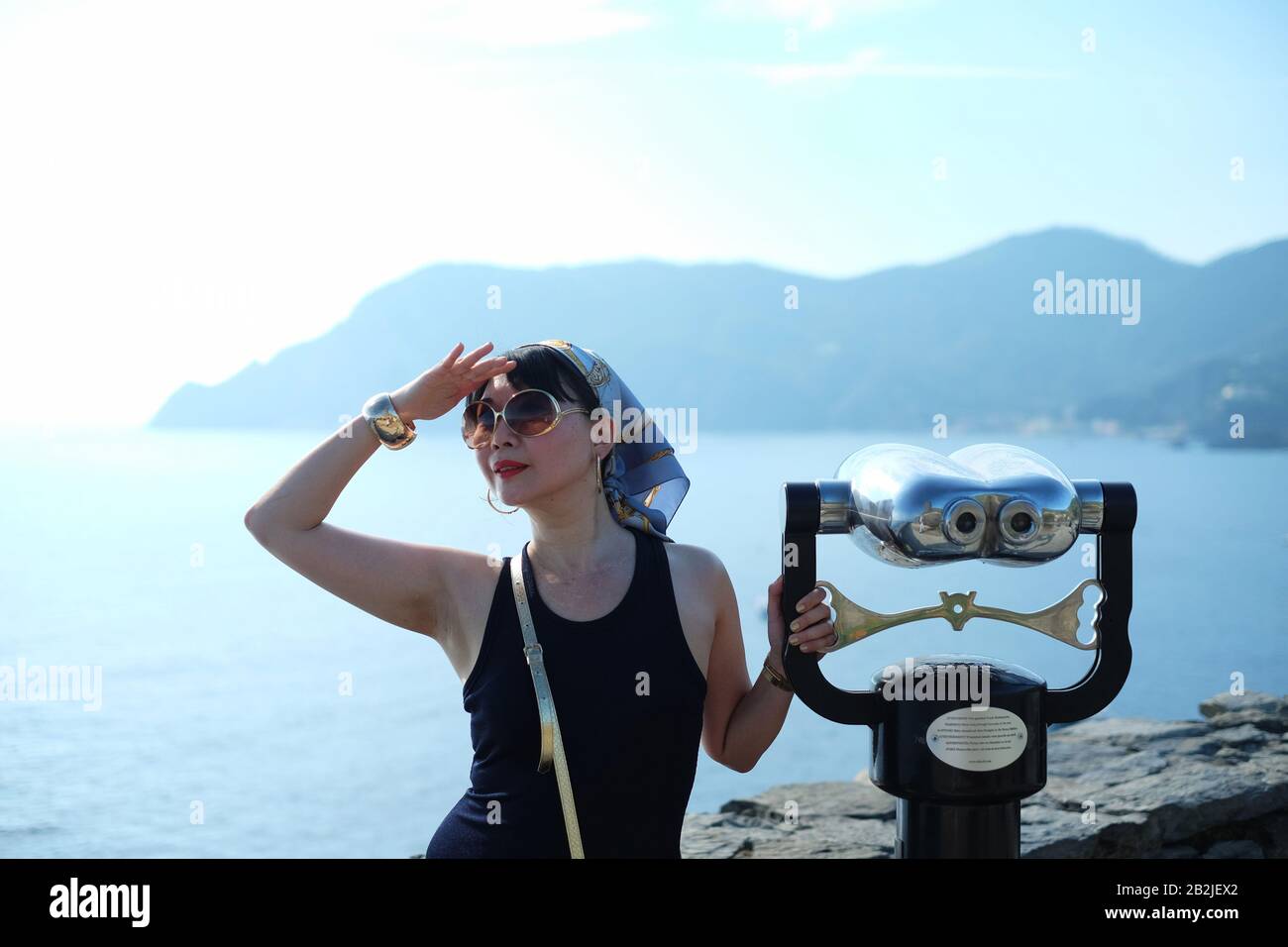 Vernazza, cinque Terre una donna glamour con velo e occhiali da sole che reggono un binocolo grande con vista panoramica sulla baia e sulle montagne di Monterosso Foto Stock
