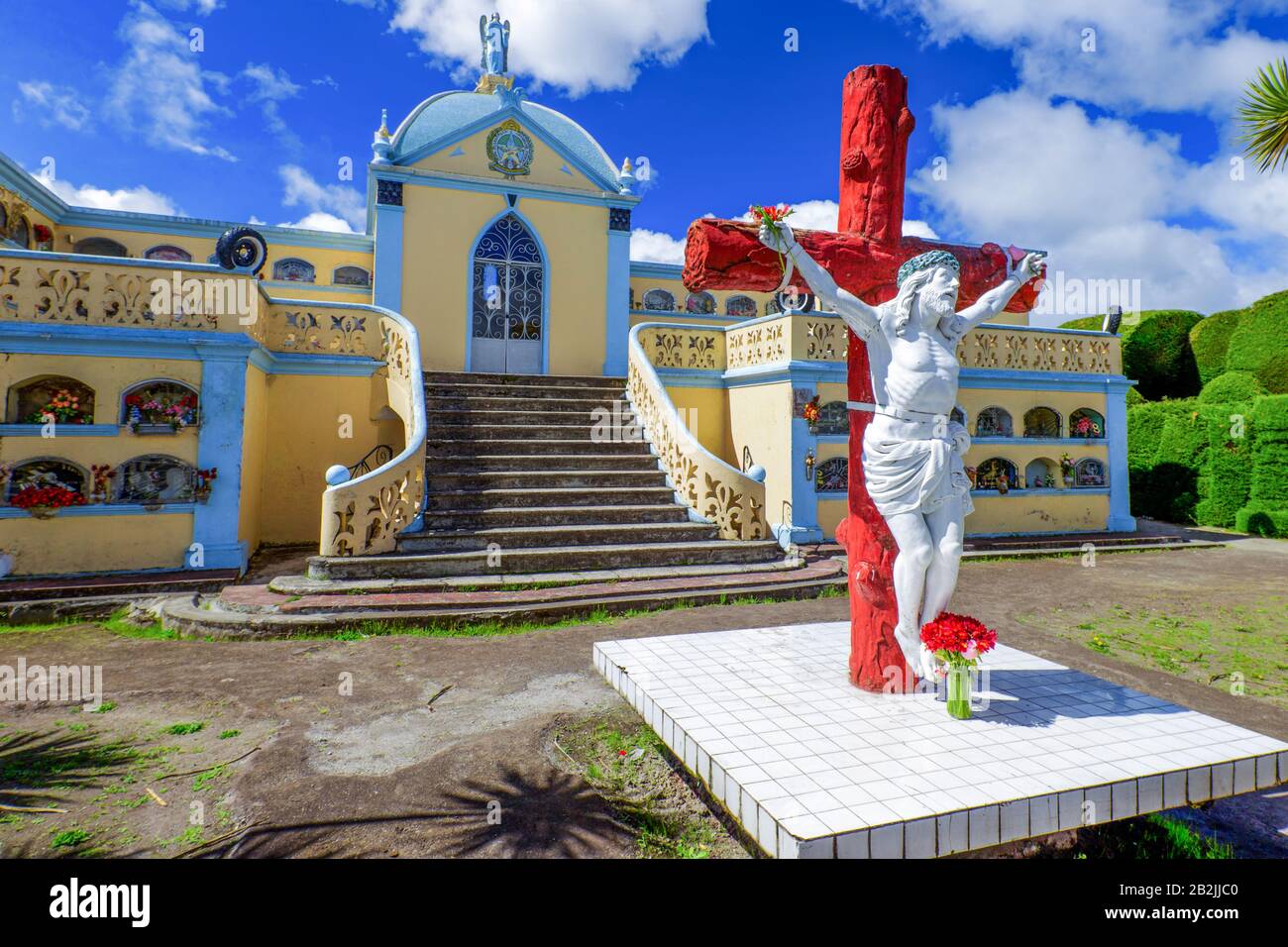 Una statua bianca di Gesù Cristo su un crocifisso rosso sorge su una piattaforma piastrellata bianca in primo piano. Dietro di essa si trova una cappella gialla e azzurra con ringhiere ornate, finestre ad arco e una centrale Foto Stock
