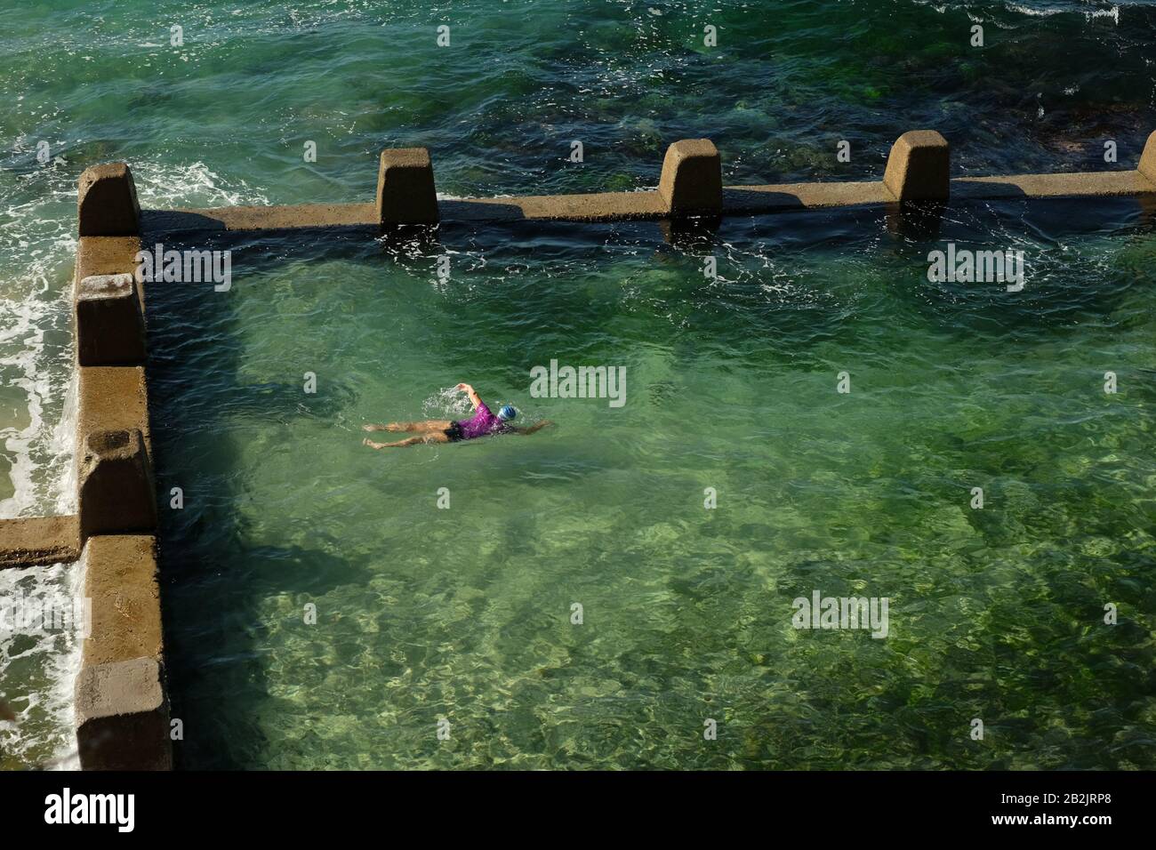 Nuotate nella fresca e verde piscina dell'oceano, l'acqua della Ross Jones Rockpool Coogee Bay, con surf che si infrangono contro le merulate mura perimetrali di Sydney Foto Stock