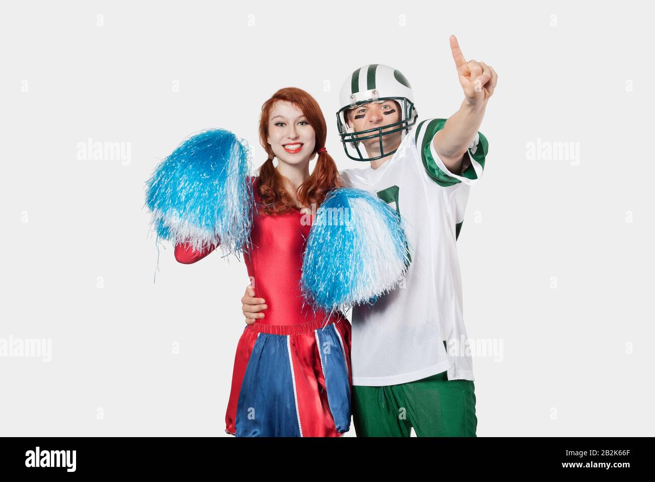 Ritratto di giovane uomo in uniforme di calcio e cheerleader con pom-pom in piedi su sfondo grigio Foto Stock