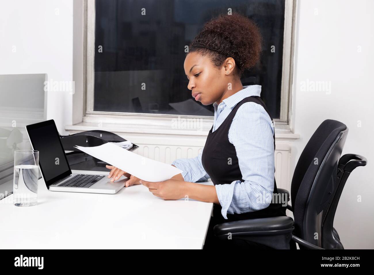 Vista laterale di una giovane donna d'affari con documenti che usano il laptop alla scrivania in ufficio Foto Stock