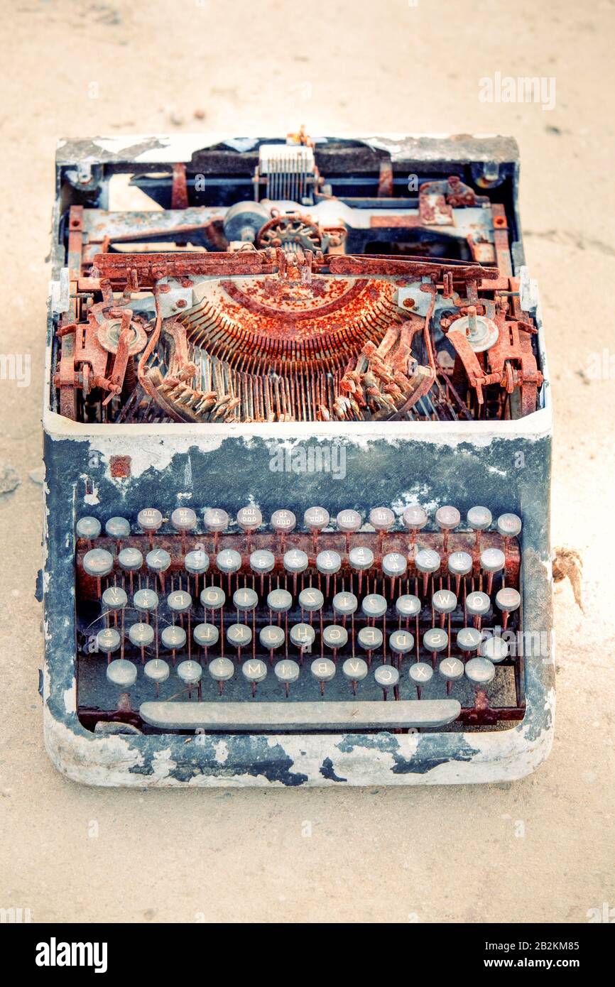 Una macchina da scrivere vintage fortemente arrugginita si trova aperta su una superficie sabbiosa, i suoi meccanismi interni esposti e corrosi. L'involucro esterno è scheggiato e staccato, rivelando strati di vernice e decomposizione. Il Foto Stock