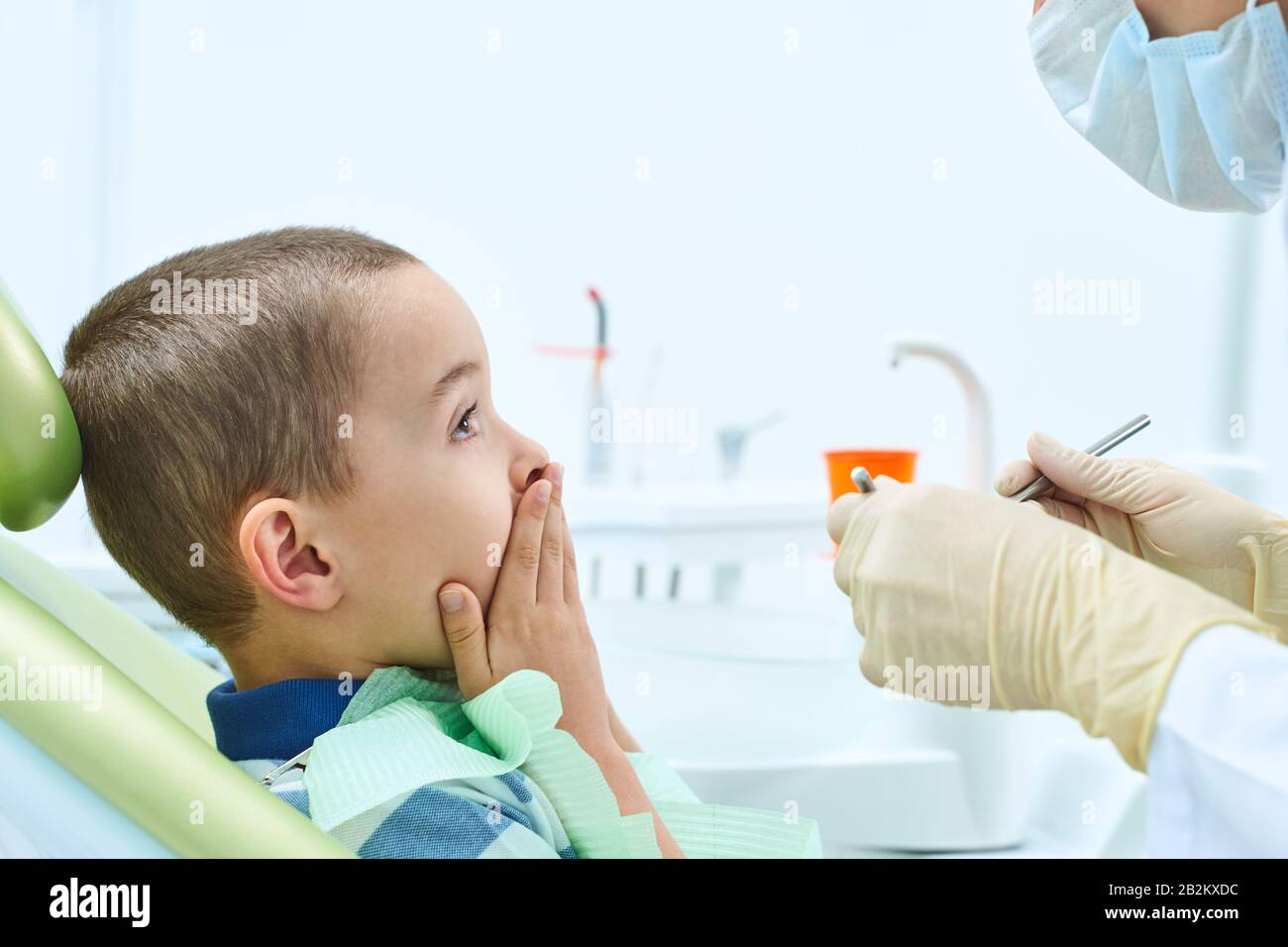 Ritratto di un bambino spaventato in una sedia dentale. Il ragazzo gli copre la bocca con le mani, impaurito di essere esaminato da un dentista. Odontoiatria dei bambini. Foto Stock