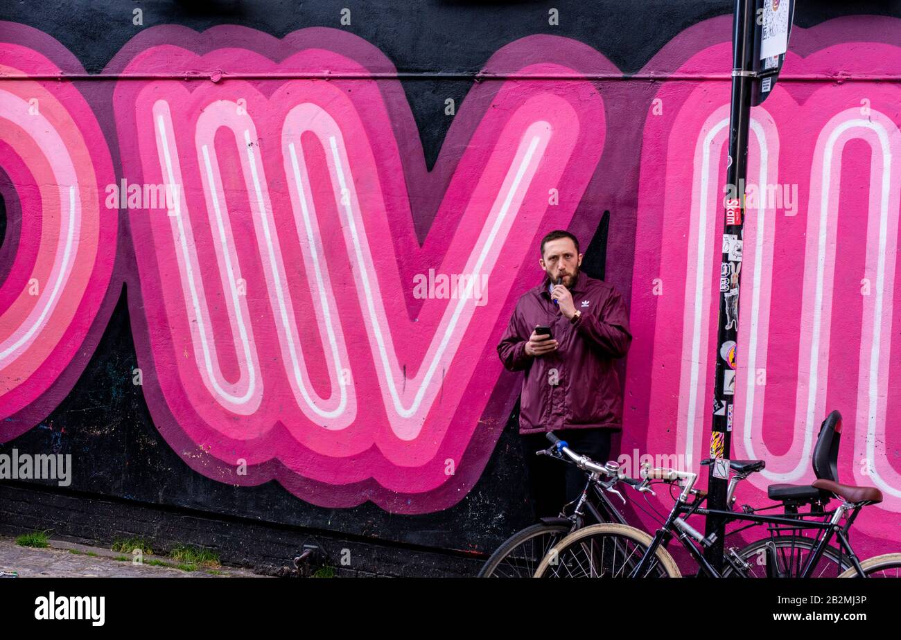 Un uomo si appoggia contro un muro con un enorme graffiti tipografico pezzo d'arte mentre vaporing, guardando il suo telefono e fissando la macchina fotografica. Foto Stock