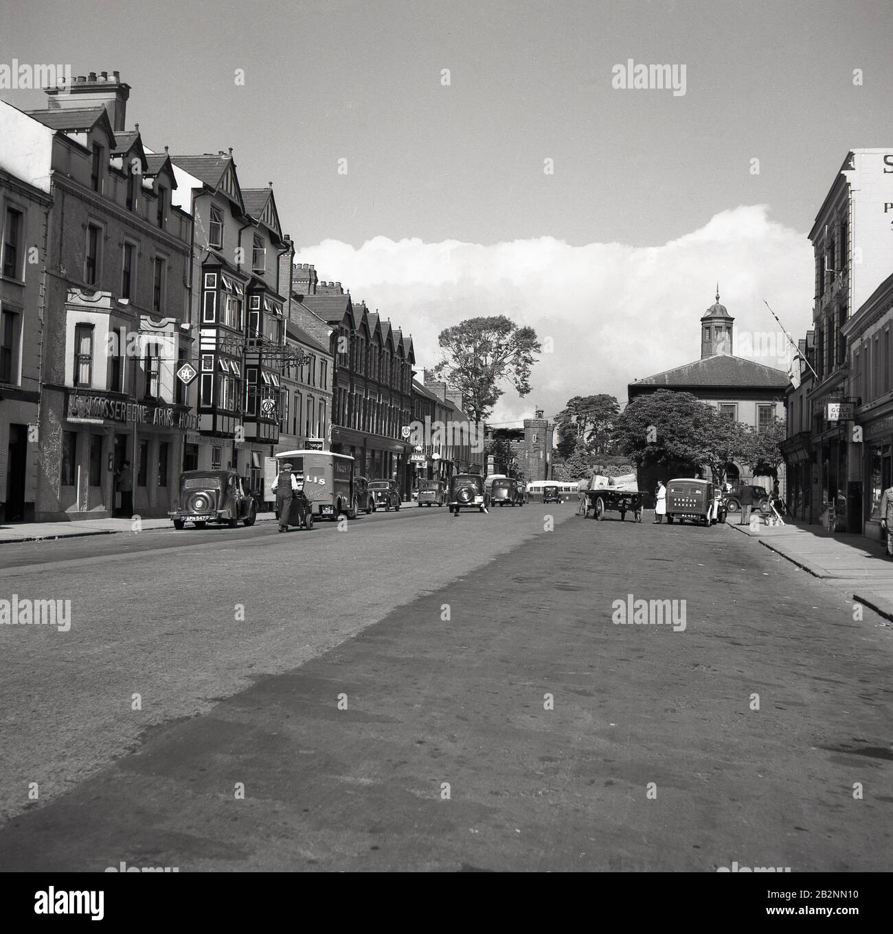 1950s, vista storica di un'ampia strada ad Antrim, Irlanda del Nord, che mostra il Massereene Arms Hotel, veicoli a motore del giorno, carrello tirato a mano e furgoni di consegna per la panetteria Ormeau (sulla destra) e la Inglis Biscuit Company (sulla sinistra) Foto Stock