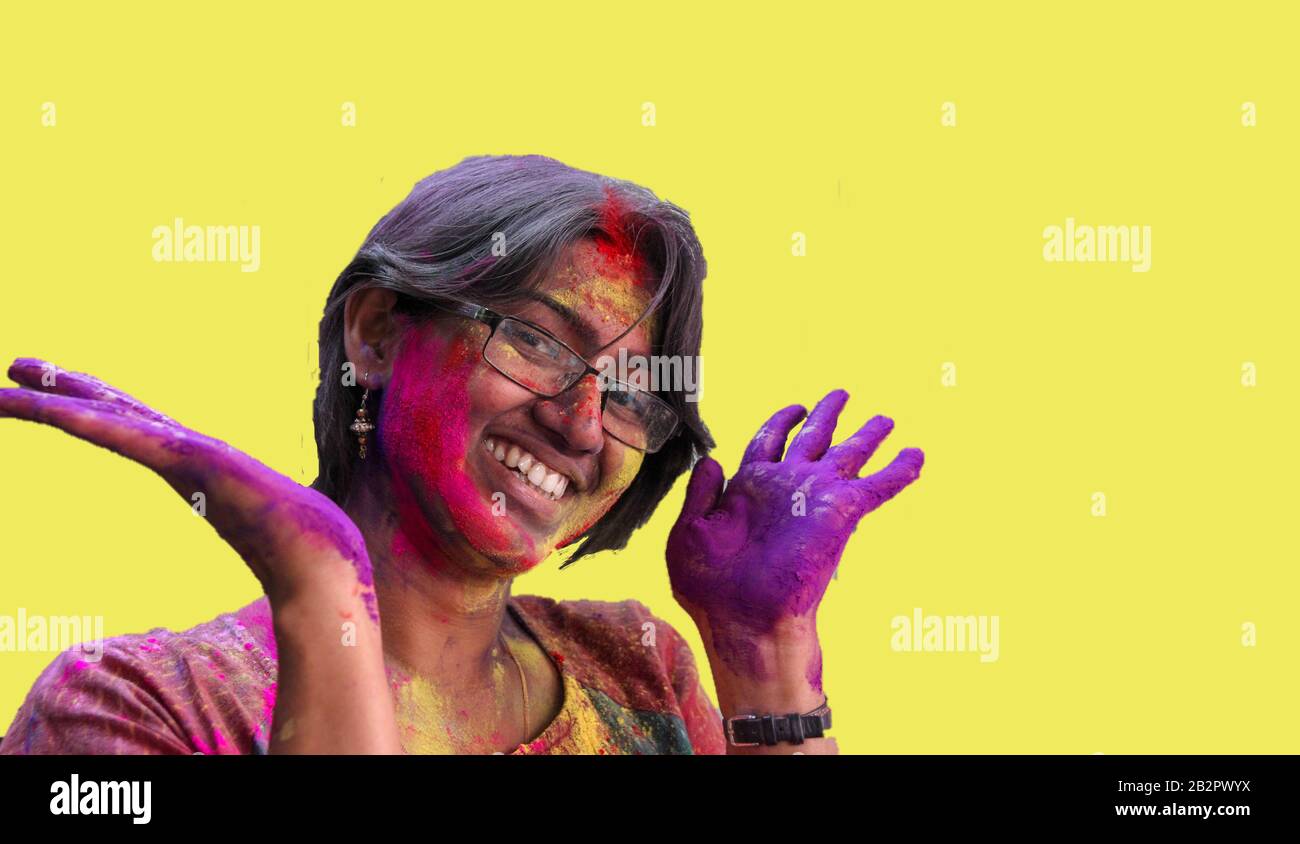 ritratto di una giovane signora con i colori holi sul viso sorridente alla fotocamera con isolato solido sfondo giallo. Foto Stock