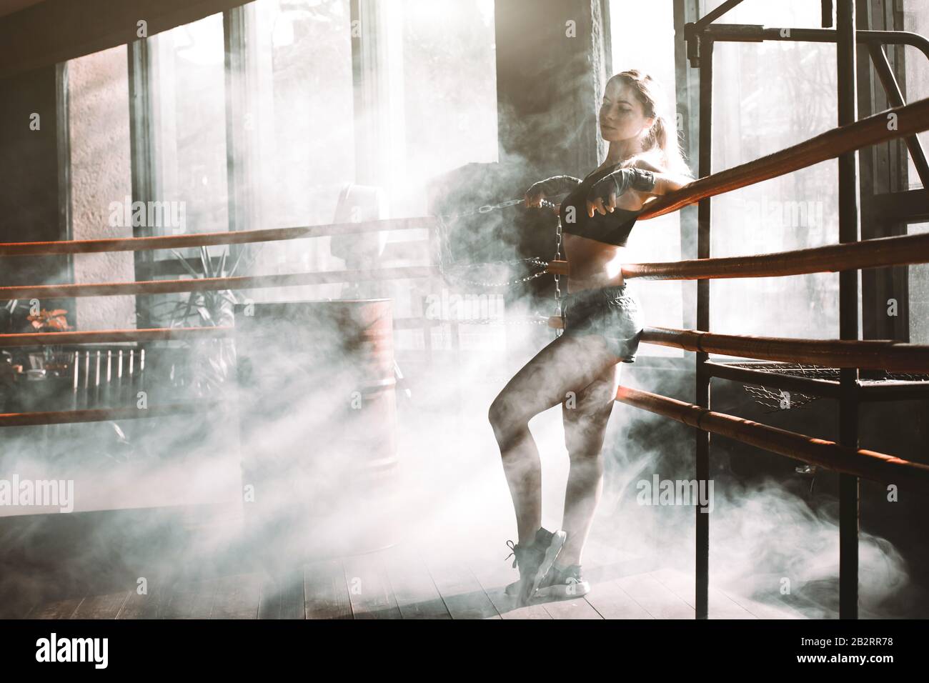 Giovane bella boxer donna in piedi sul ring. Ritratto completo del corpo di donna boxer che indossa reggiseno sportivo nero, pantaloni grigi, allenatori in piedi in anello e. Foto Stock