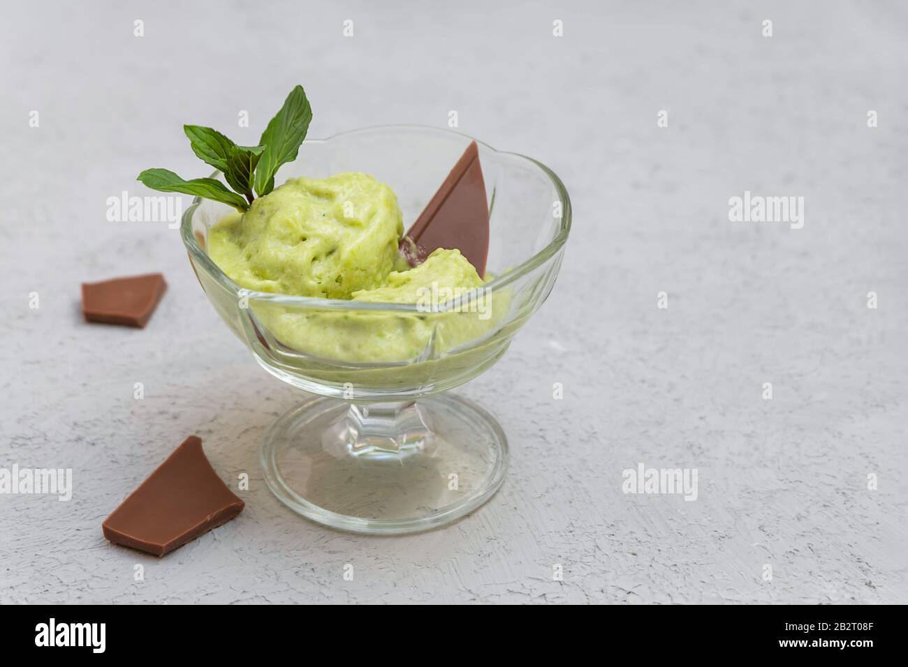 Ciotola di vetro con gelato avocado, foglie di menta e pezzi di cioccolato su sfondo grigio neutro con spazio di copia Foto Stock