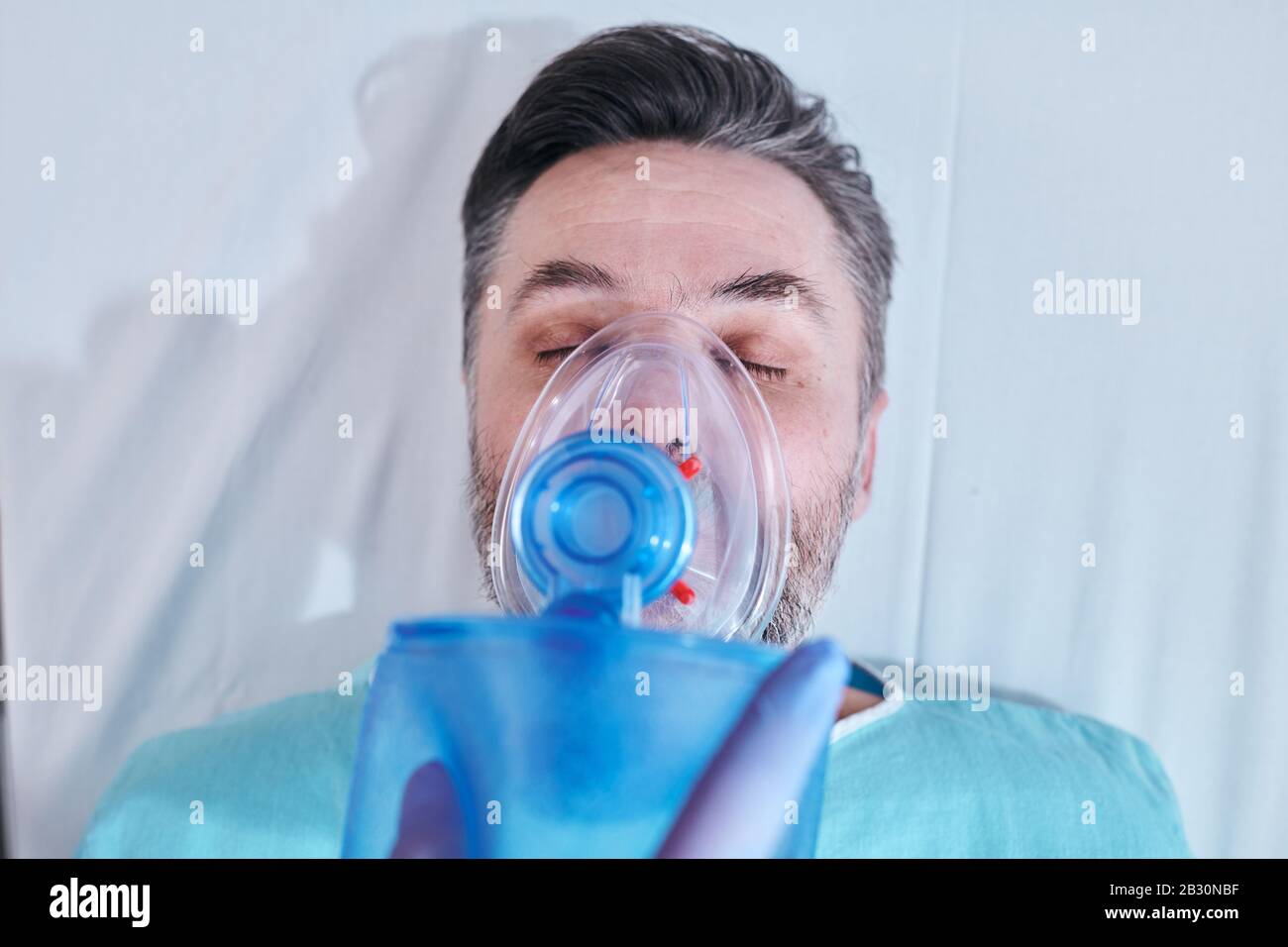 Vista dall'alto del paziente maturo in condizioni gravi che mantiene gli occhi chiusi durante l'inalazione di ossigeno attraverso la maschera della valvola della sacca Foto Stock