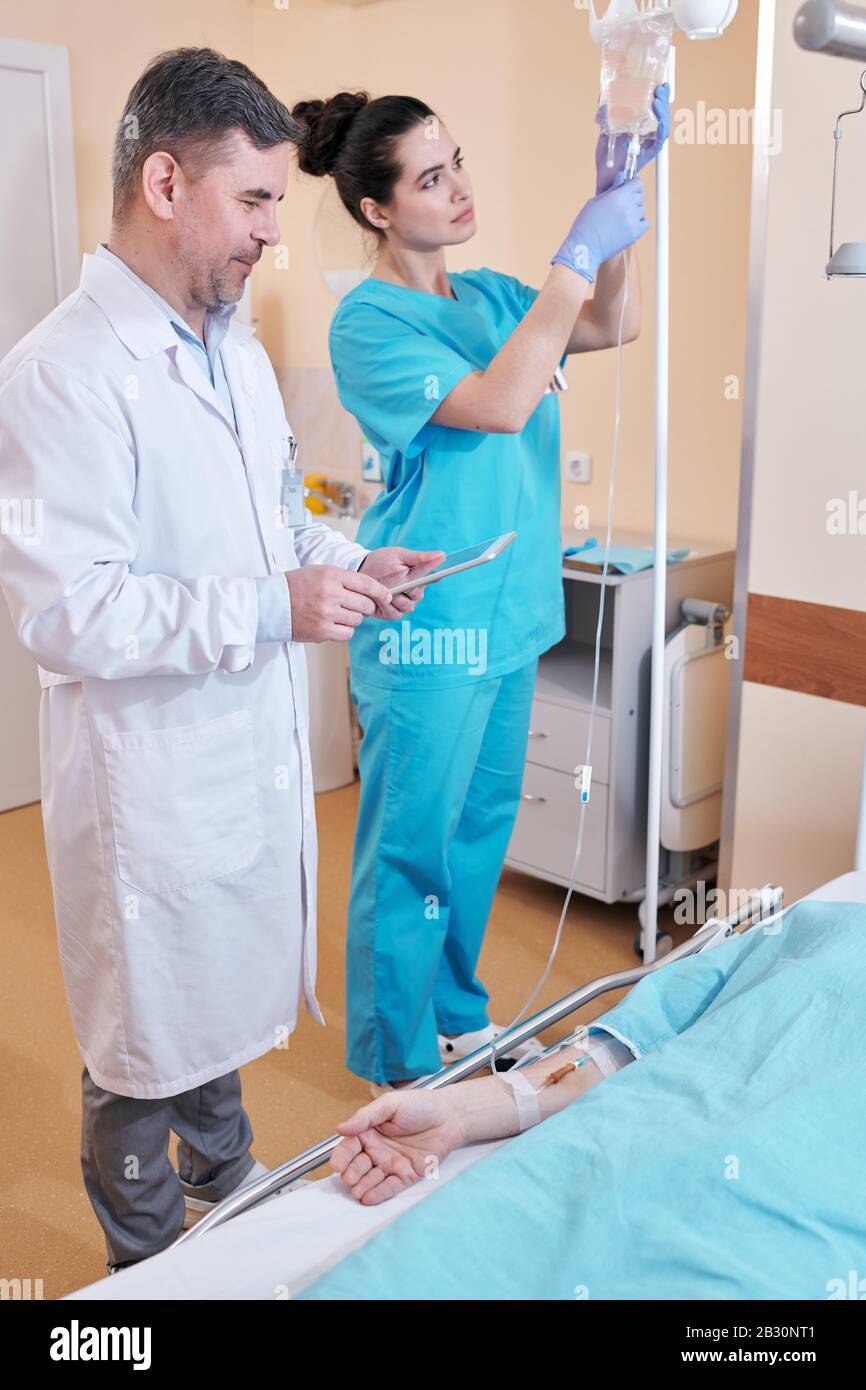 Infermiere giovane che prepara la sacca per infusione endovenosa mentre dà al paziente IV, il medico maturo che parla con il paziente durante la visualizzazione dei risultati del test sulla compressa Foto Stock