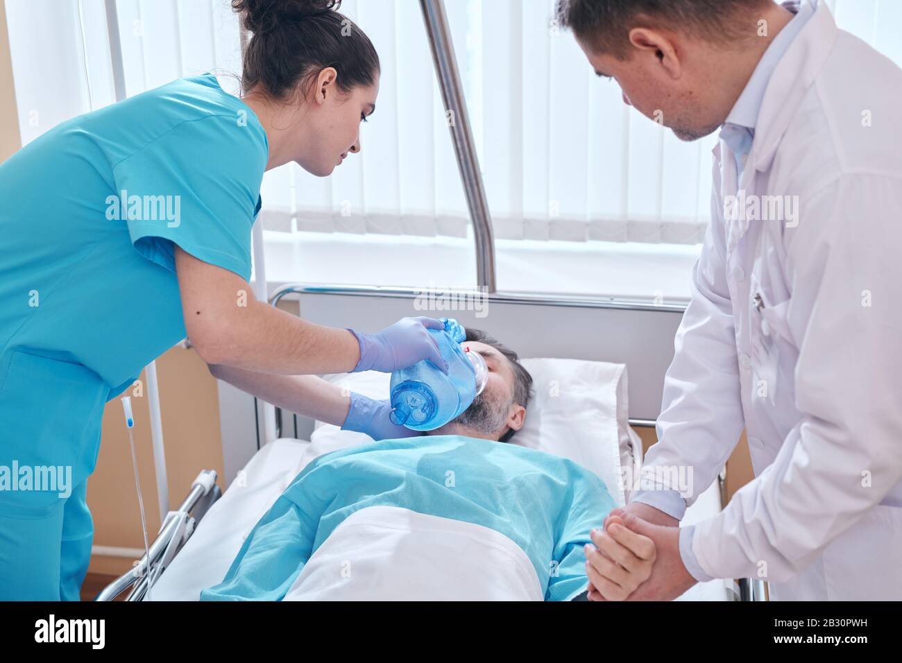 Giovane infermiera che mette maschera di ossigeno della valvola della sacca ai pazienti faccia e medico che controlla il suo impulso mentre forniscono assistenza urgente all'uomo Foto Stock