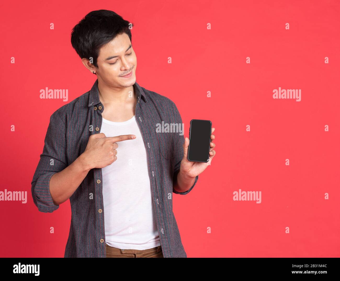 Ritratto di giovane e attraente uomo d'affari asiatico o studente utilizzando telefono cellulare, laptop, tablet, in piedi isolato su sfondo rosso indossando camicia casual Foto Stock