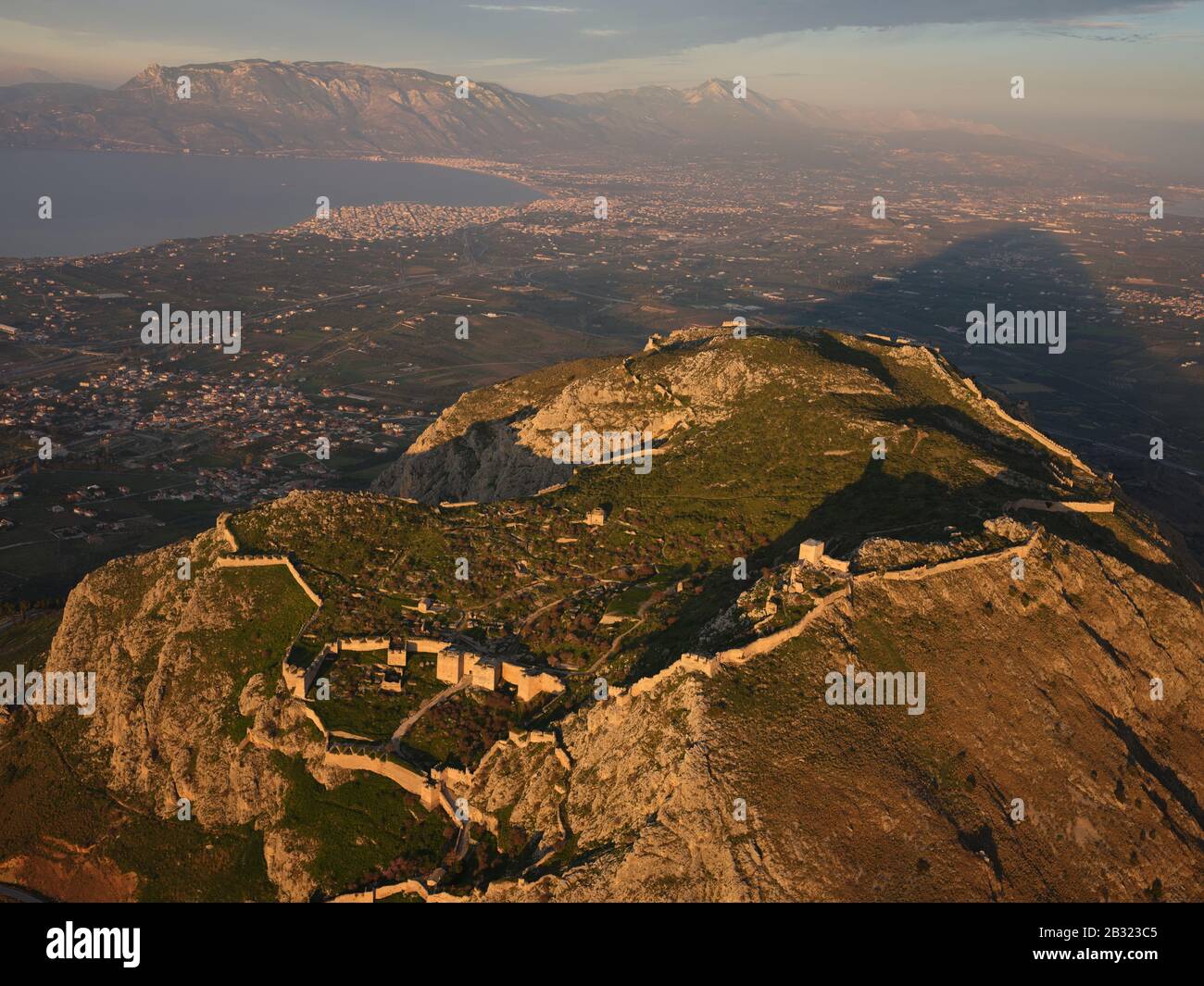 VISTA AEREA. Antica città di Corinto che domina il Golfo di Corinto e la nuova città di Corinto al tramonto. Peloponneso, Grecia. Foto Stock