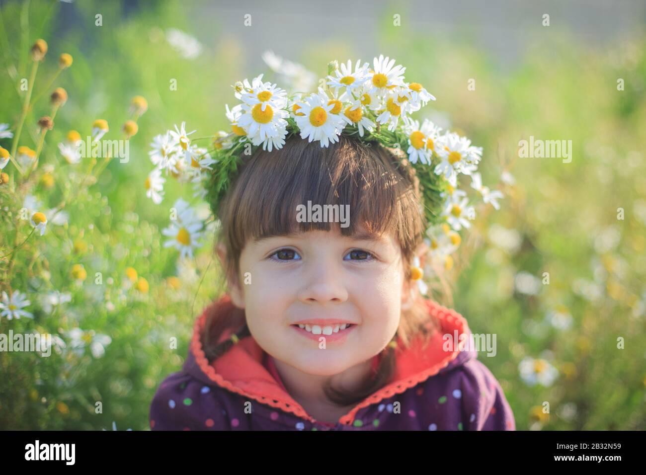Una bambina in una corona di margherite sorride e si siede nell'erba verde in estate. Giornata di sole. Foto Stock