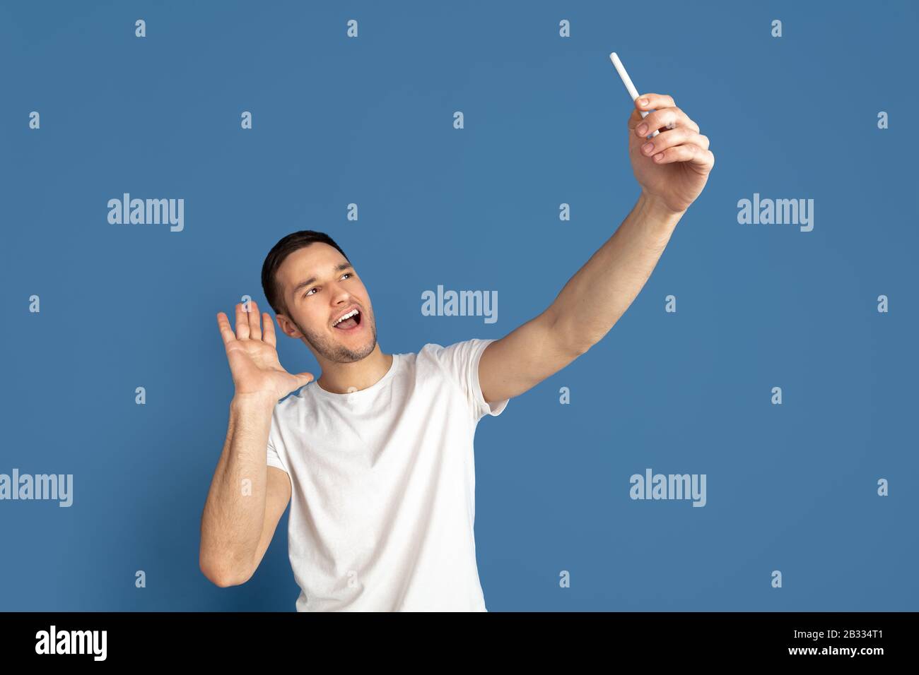 Prendere selfie. Ritratto del giovane caucasico isolato su sfondo blu studio. Bel modello maschile in stile casual, colori pastello. Concetto di emozioni umane, espressione facciale, vendite, pubblicità. Foto Stock