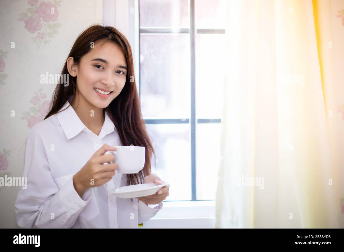 Donne asiatiche a bere caffè e svegliarsi nel suo letto pienamente riposati e aprire le tende al mattino per ottenere aria fresca sul sole Foto Stock