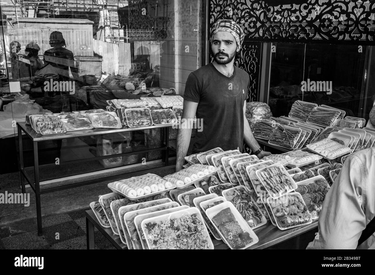 Aqaba, GIORDANIA - 31 GENNAIO 2020: Giovane uomo con barba e sciarpa a testa araba vende dolci fuori strada nella soleggiata giornata invernale, mercato pubblico della città. Immagine in bianco e nero Foto Stock