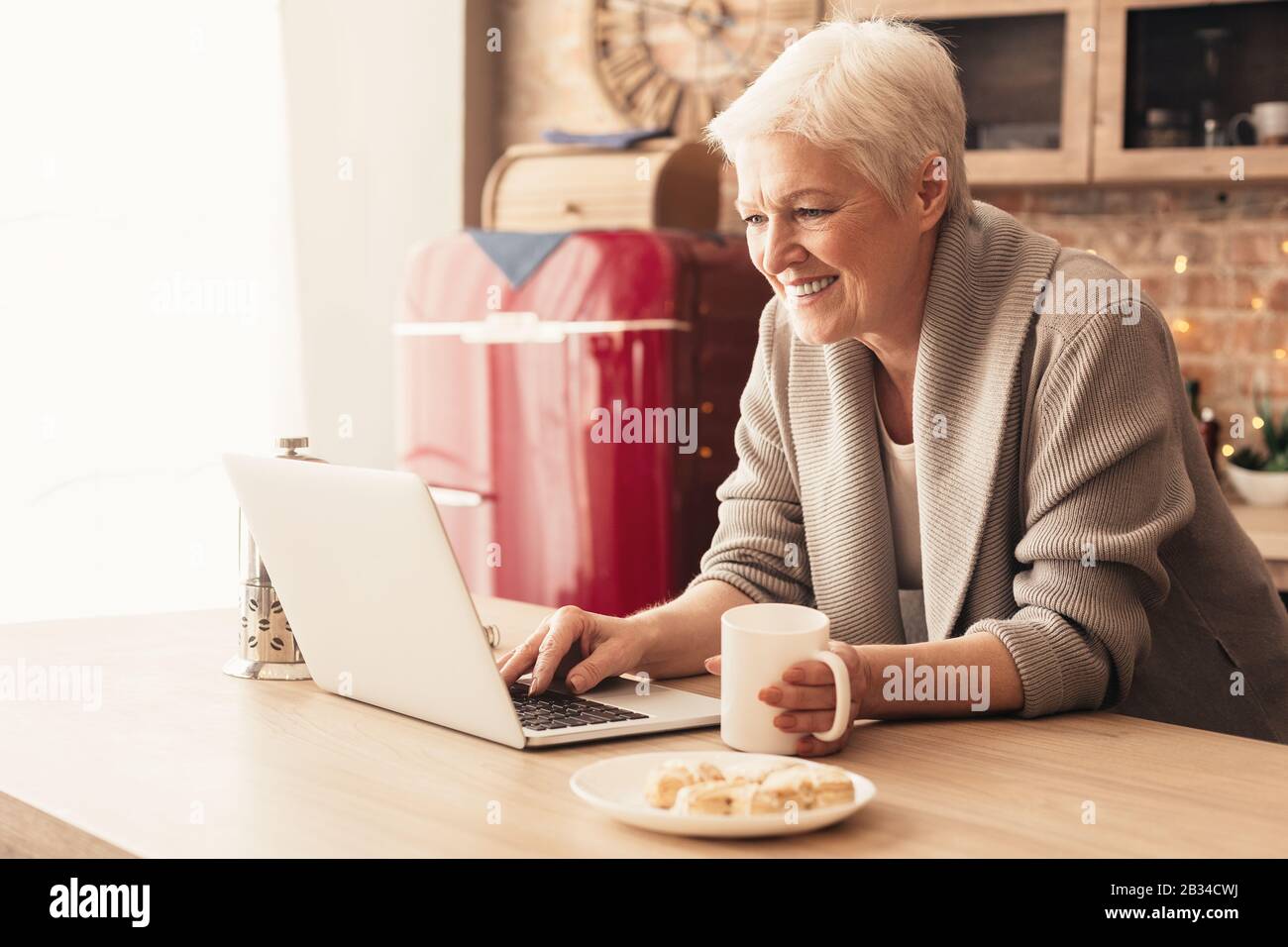 Donna Sorridente In Età Rilassante In Cucina Con Laptop E Tè Foto Stock