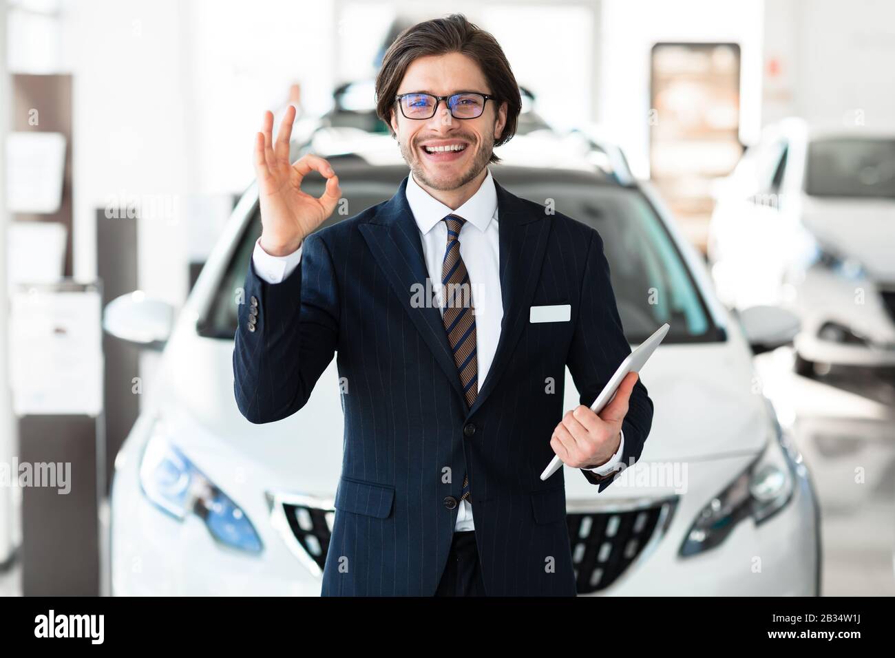Sorridente Salesman Gesturing Okay Standing In Luxury Auto Store Foto Stock