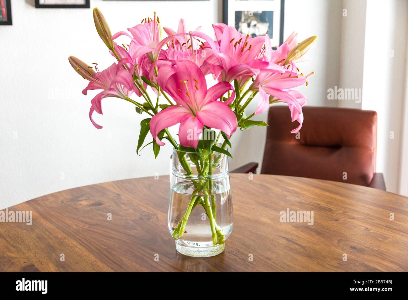 Bella gigli rosa fresco con una brocca di vetro come vaso su un bel tavolo di acacia in legno con una poltrona in pelle di cognac contro una parete bianca con... Foto Stock