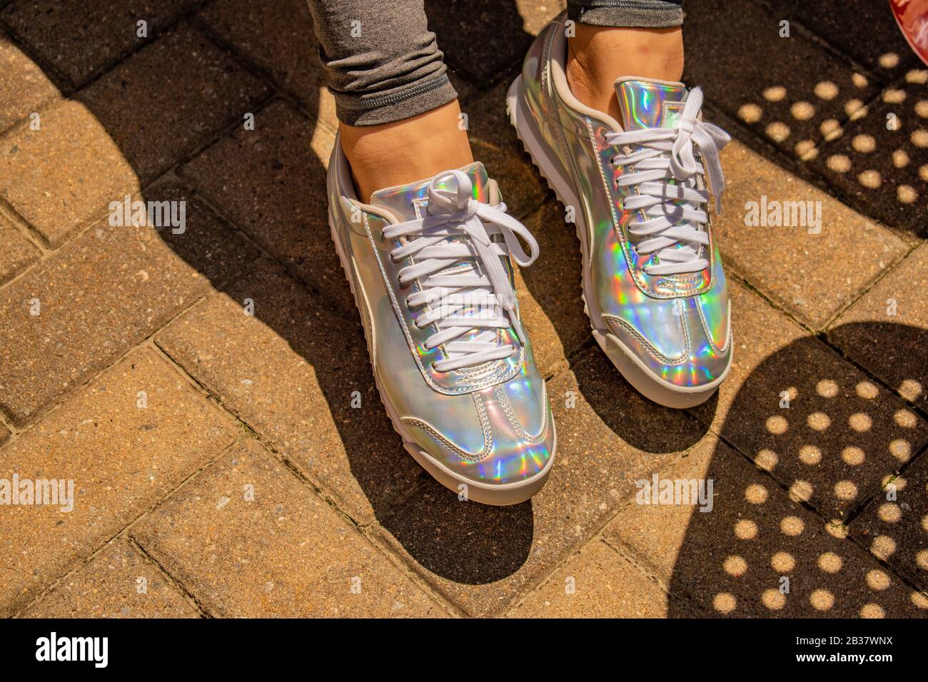 Due piedi femminili con sneakers iridescenti colorate in luce del sole che creano ombre sulle finitrici in mattoni. Foto Stock