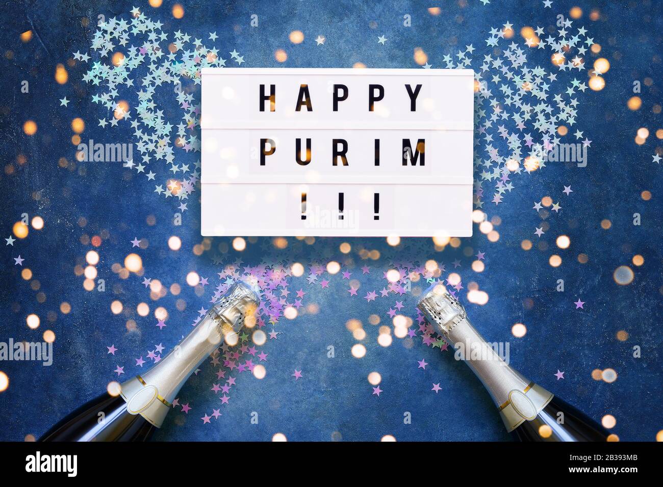 Happy Purim scritto in scatola leggera e due bottiglie di champagne con confetti olografici stelle su sfondo blu. Piatto di concetto di celebrazione Purim. Foto Stock