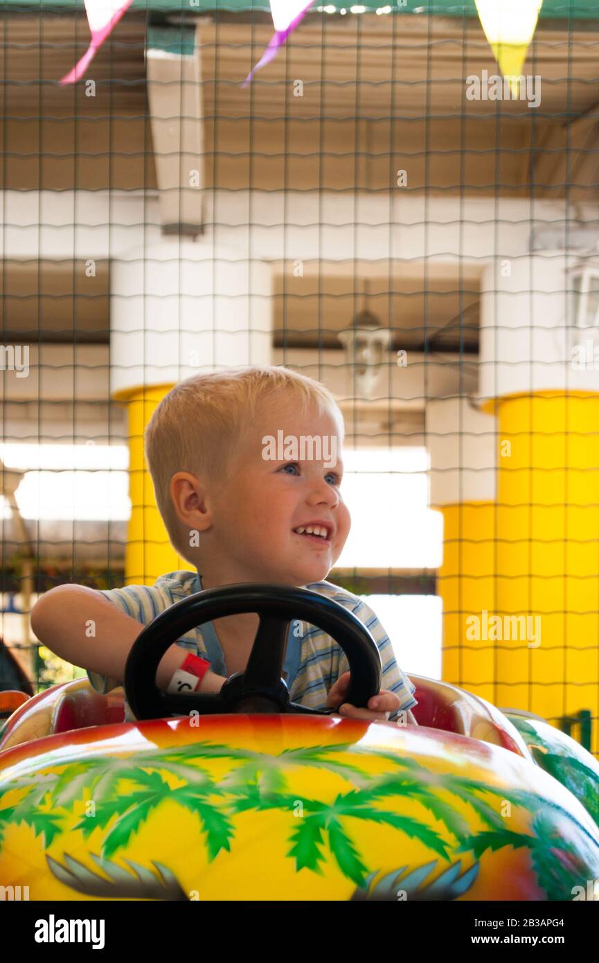 Un ragazzino biondo molto gioioso e felice di tre anni è in sella a un'auto gialla con un'auto verde nel parco divertimenti estivo Riviera, Sochi con la schiena allo spettatore. Riposo, tropici, riposo, attivazione, sorriso. Foto verticale Foto Stock