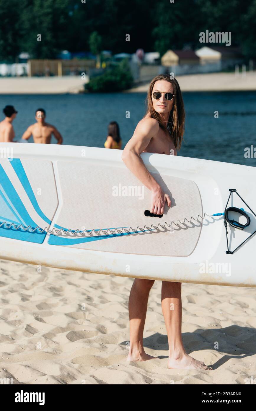 bel giovane uomo in occhiali da sole che tiene surf board e guardare lontano sulla spiaggia Foto Stock