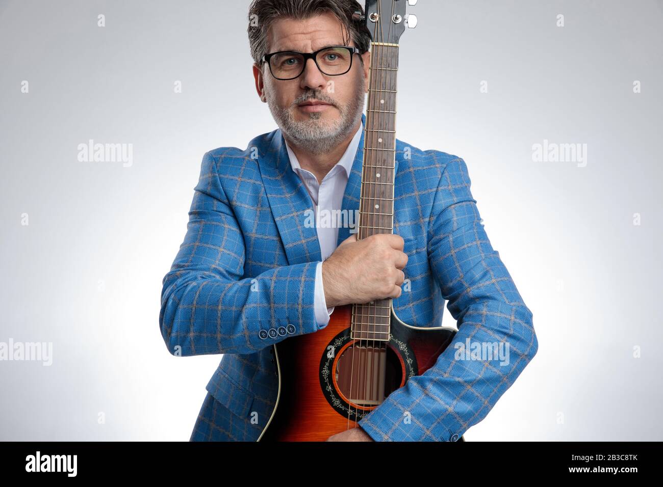 ritratto di un affascinante uomo d'affari formale indossando un vestito azzurro e occhiali in piedi e tenendo la sua chitarra stretta mentre guarda la fotocamera seri Foto Stock