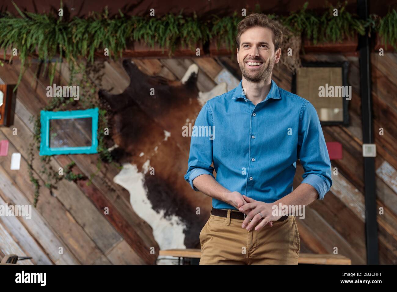 L'uomo positivo casual sfrega le mani, sorridendo e guardando lontano  indossando una camicia blu e pantaloni marroni, in piedi su uno sfondo di  coffee shop Foto stock - Alamy