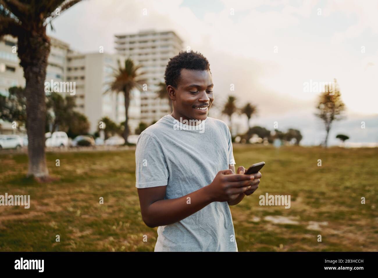 Ritratto di un giovane atleta sorridente in piedi nel parco al mattino SMS messaggi sul telefono cellulare hi Foto Stock