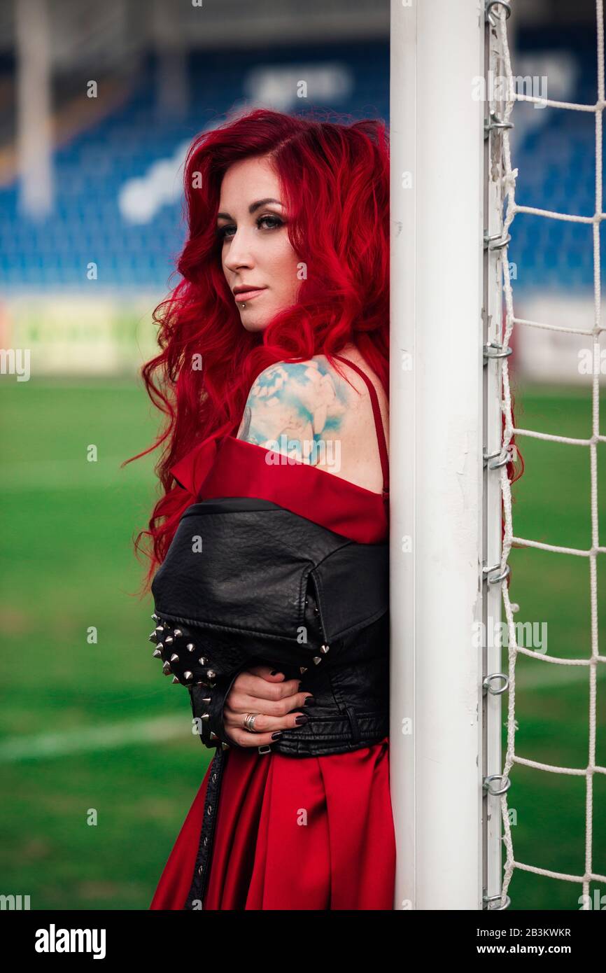 Donna lussuosa con capelli rossi e in un abito rosso gioca sul campo da calcio. Idea e concetto di una combinazione di sport e bellezza, presentazione inusuale Foto Stock