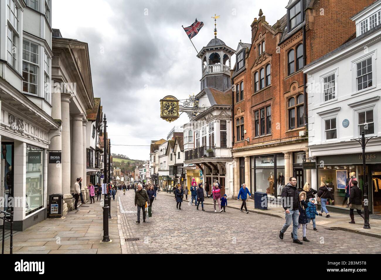 Gli amanti dello shopping a Guildford High Street passano accanto alla Guildhall con il suo famoso orologio risalente al 1693. Sotto i piedi si trovano le pietre di granito di Guildford. Foto Stock