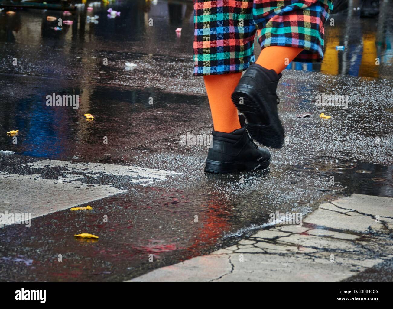 Gambe con pantaloni colorati controllati, calzini arancioni luminosi e calze nere che si schiaccia su asfalto grigio scuro bagnato Foto Stock