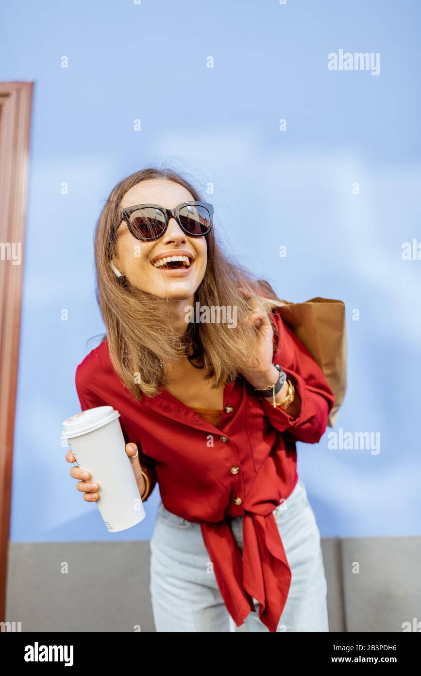 Stile di vita ritratto di una donna elegante in camicia rossa in piedi con tazza da caffè e borsa all'aperto sul colore sfondo parete. Caffè da asporto e stile donna Foto Stock