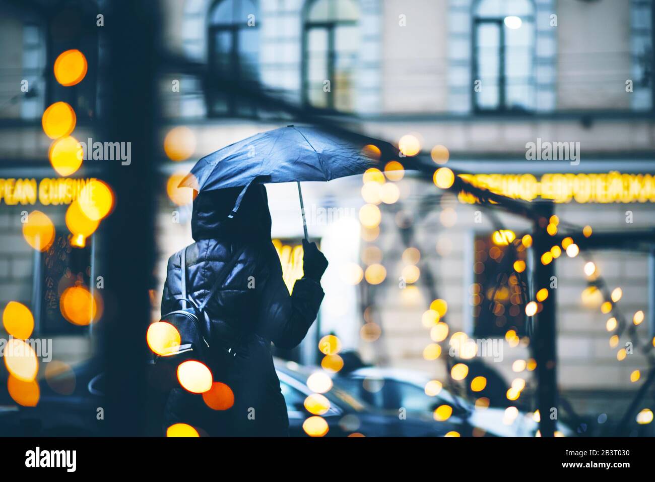 Una ragazza in un frangivento nero con un ombrello bagnato cammina lungo una strada della città in serata, illuminato da luci che decorano la recinzione nera. Foto Stock