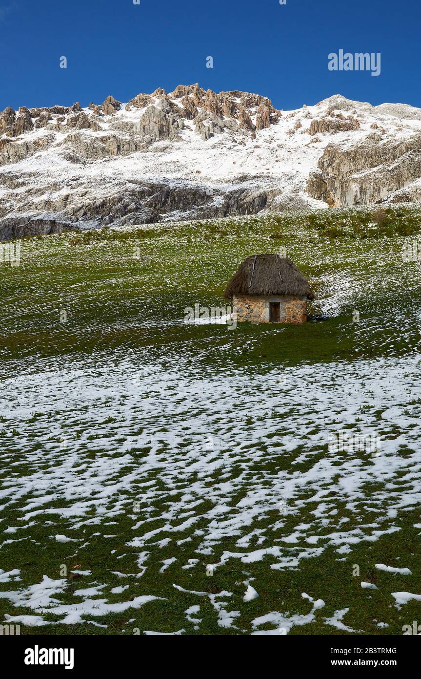 Teito, tradizionale dimora in pietra con tetto di paglia, con montagne innevate sullo sfondo (Valle del Lago, Parco Naturale Somiedo, Asturie, Spagna) Foto Stock