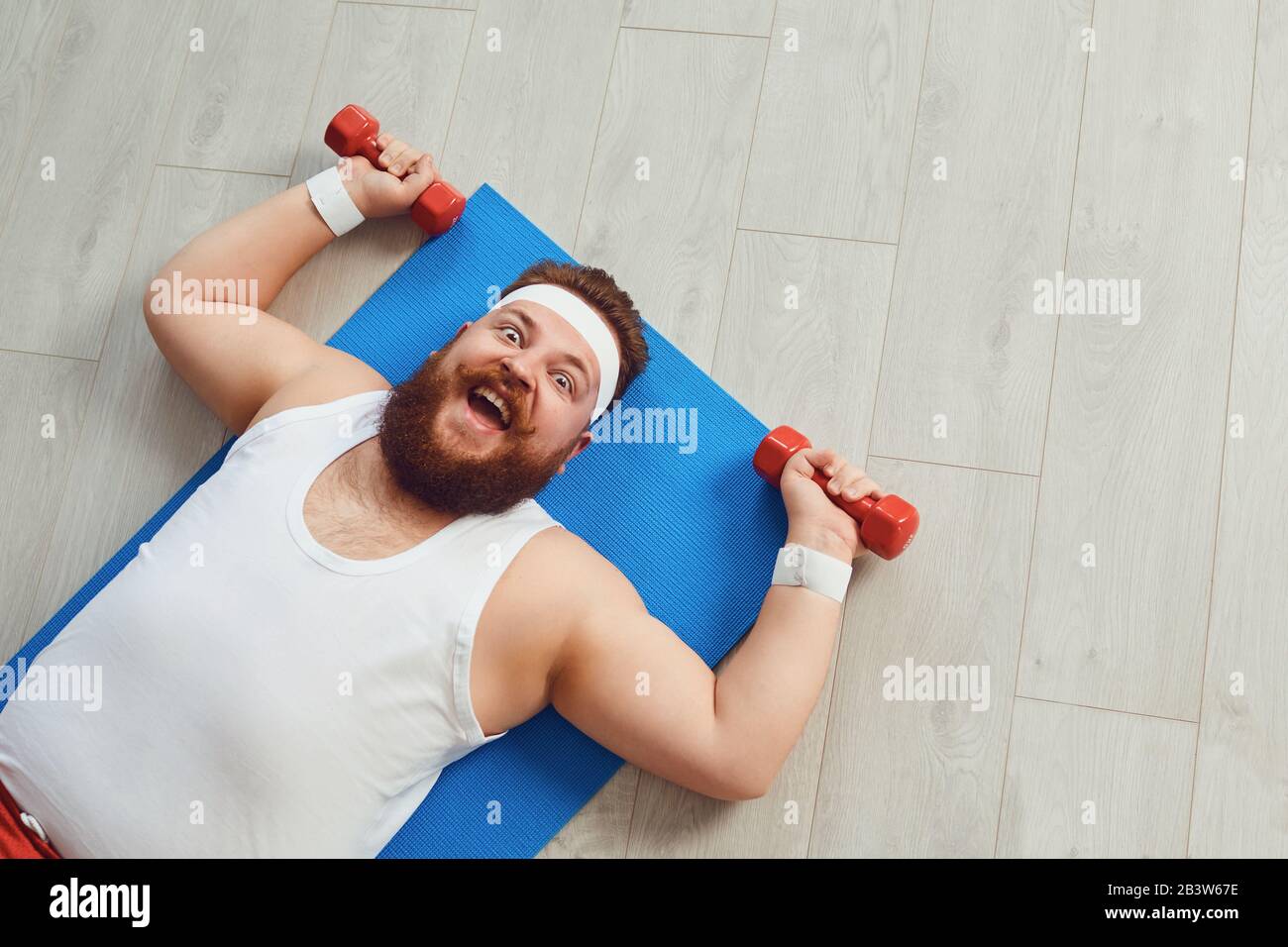Divertente uomo grasso fare esercizi con dumbbells su un allenamento sul pavimento in casa. Foto Stock