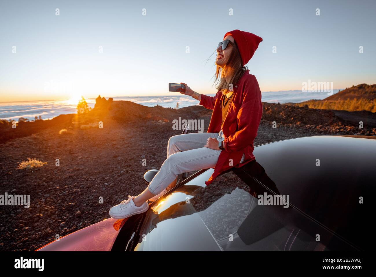 Giovane donna vestita di rosso che gode il paesaggio del tramonto sopra le nuvole, seduto sul tetto della macchina durante il viaggio in montagna. Stile di vita spensierato e concetto di viaggio Foto Stock