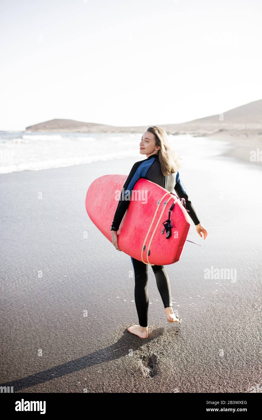 Giovane donna in muta a piedi con tavola da surf, lasciando impronte sulla sabbia dietro, vista dal retro. Sport acquatici e concetto di stile di vita attivo Foto Stock