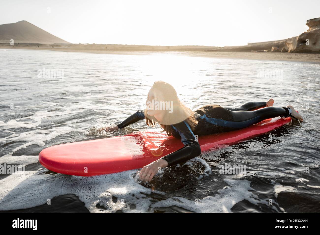 Giovane donna nella muta che cattura il flusso d'acqua sulla tavola da surf, surf sull'oceano ondulato durante un tramonto. Sport acquatici e concetto di stile di vita attivo Foto Stock