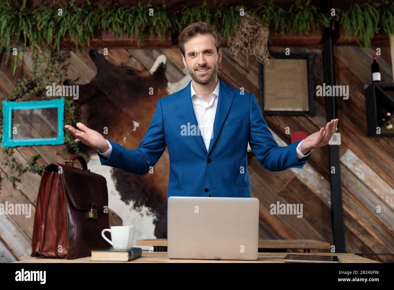 Allegro uomo d'affari sorridente e invitante con le mani aperte mentre si sta dietro una scrivania con un laptop, valigetta, tazza da caffè, tablet e un boo Foto Stock