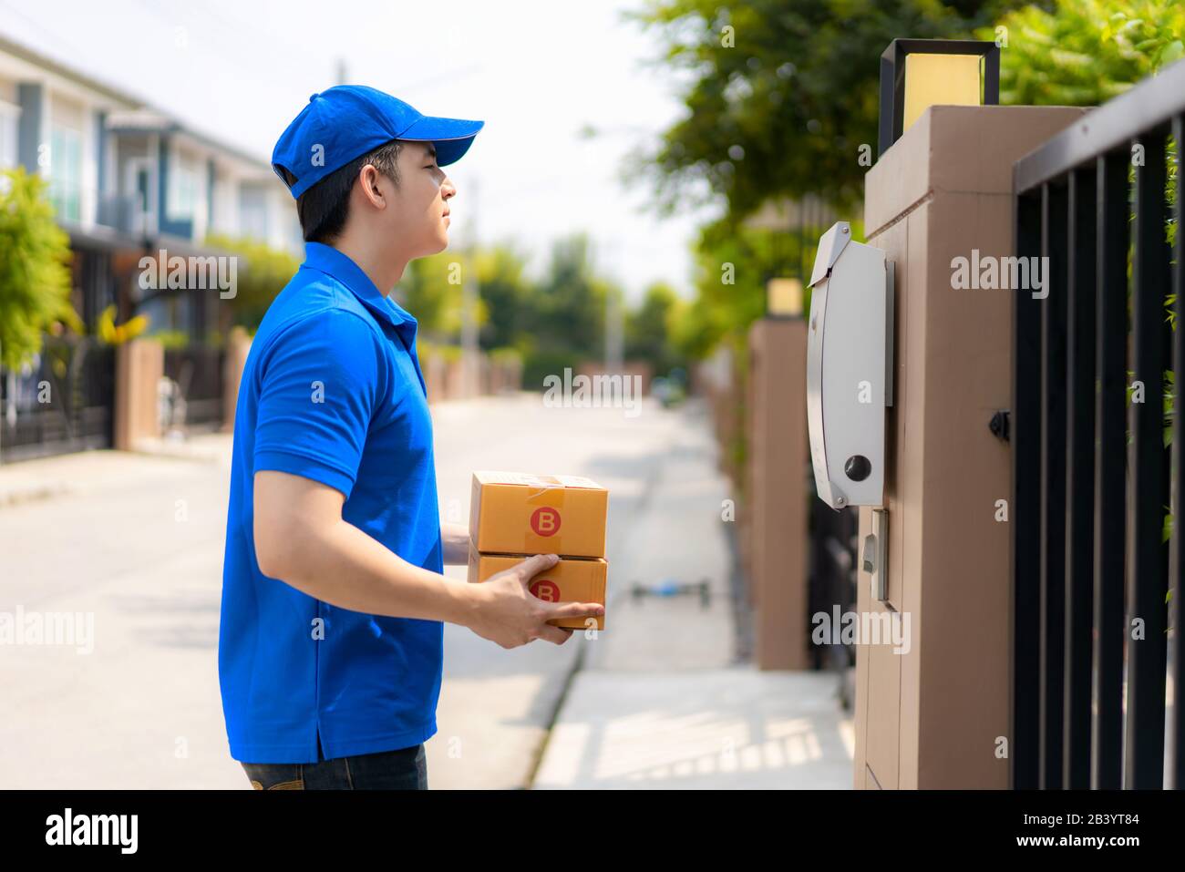 Asian consegna giovane uomo in blu uniforme sorriso e tenendo mucchio di scatole di cartone in casa villaggio di fronte con spazio copia. Pubblicità, Business, Tran Foto Stock