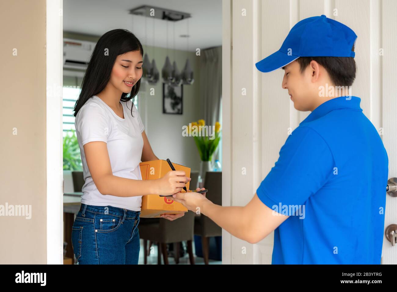Il giovane uomo asiatico della consegna in un sorriso blu uniforme e tenendo lo smartphone davanti a casa e la donna asiatica firma nello smartphone ad accettare un deliv Foto Stock