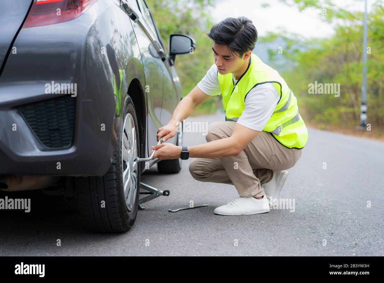 Giovane uomo asiatico con giubbotto di sicurezza verde che sostituisce il pneumatico forato sulla sua auto allentando i dadi con una chiave per ruota prima di sollevare il veicolo Foto Stock