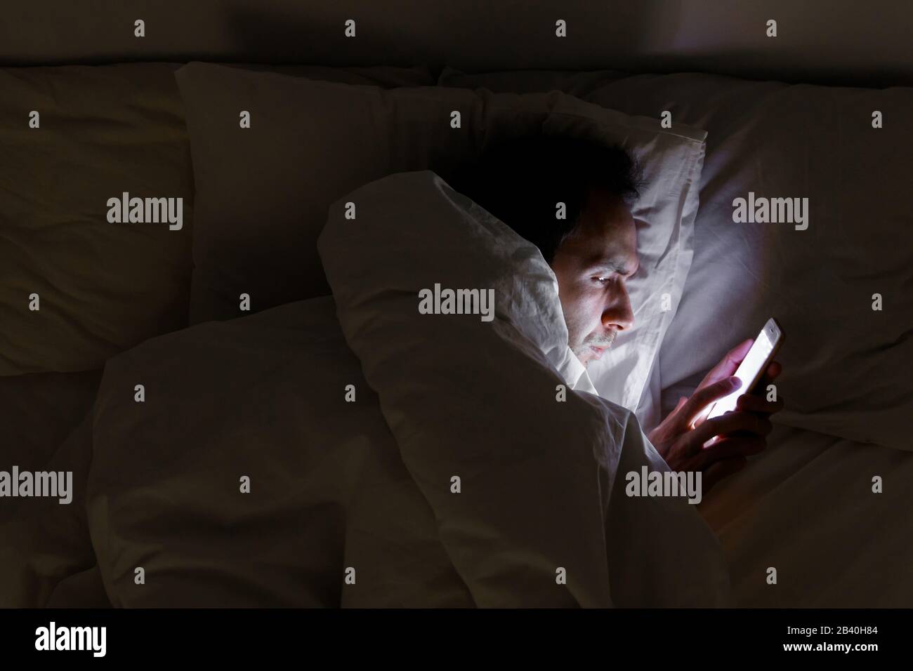 Dipendenza mobile, nomofobia, insonnia, concetto di disturbo del sonno. L'uomo annoiato che chiacchierava e naviga su internet con lo smartphone in tarda notte, sdraiato su BE Foto Stock