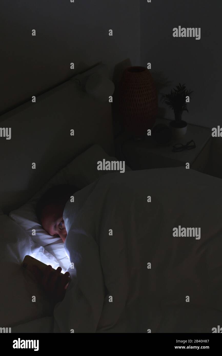 Dipendenza mobile, nomofobia, insonnia, concetto di disturbo del sonno. L'uomo annoiato che chiacchierava e naviga su internet con lo smartphone in tarda notte, sdraiato su BE Foto Stock