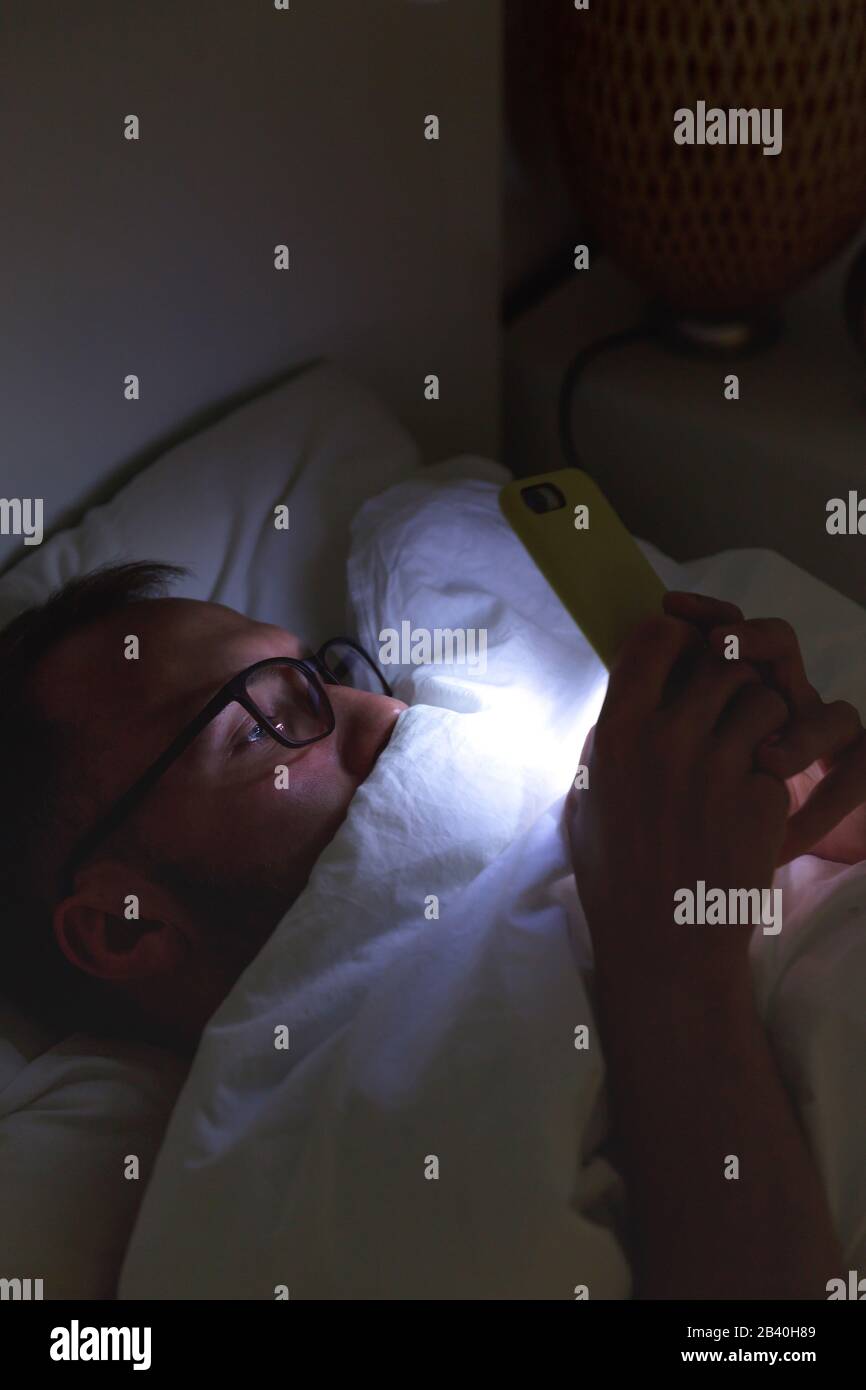 Dipendenza, nomofobia, insonnia. Un primo piano di un uomo assonnato in occhiali utilizzando lo smartphone, sdraiato a letto sotto la coperta a tarda notte, non può dormire. Foto Stock
