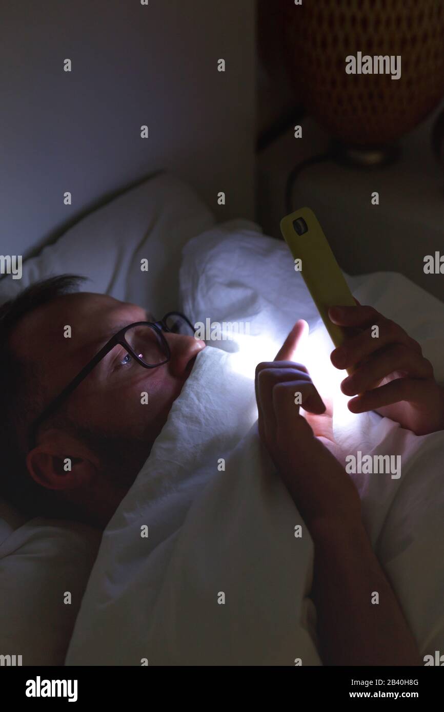 Dipendenza, nomofobia, insonnia. Un primo piano di un uomo assonnato in occhiali utilizzando lo smartphone, sdraiato a letto sotto la coperta a tarda notte, non può dormire. Foto Stock